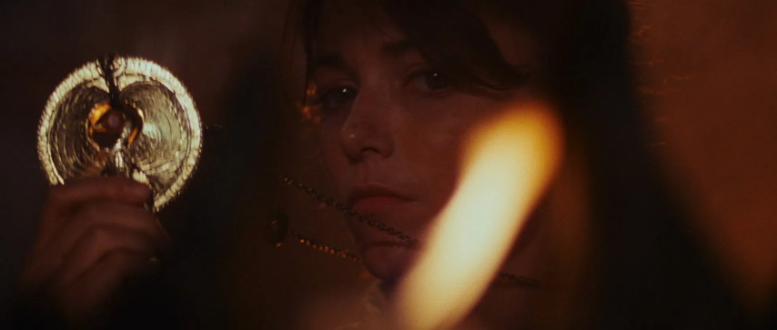 An image from the movie "Raiders Of The Lost Ark". Marion holds up a golden medallion, lit by a candle's flame.