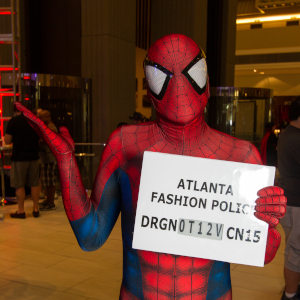 A photograph of a person in a Spider-Man costume. He holds a board that reads 'Atlanta Fashion Police' with one hand and shrugs with the other hand