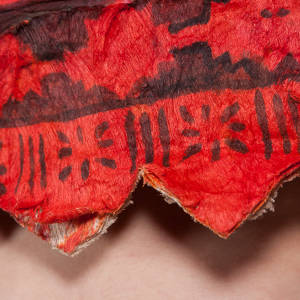 An extreme closeup of red fabric marked with dark dye in intricate patterns. The zig-zag edge of the fabric reveals skin beneath.