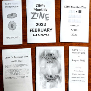 A photograph of several zines layed out on a wooden table. Each says Cliff's Monthly Zine and each has a different date.